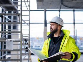 A man in a hard hat is holding a binder and is looking out into the distance while he’s standing in a construction zone.