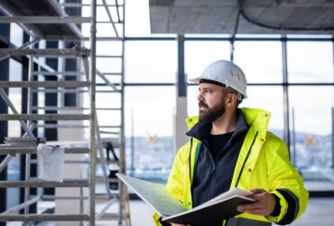 A man in a hard hat is holding a binder and is looking out into the distance while he’s standing in a construction zone.