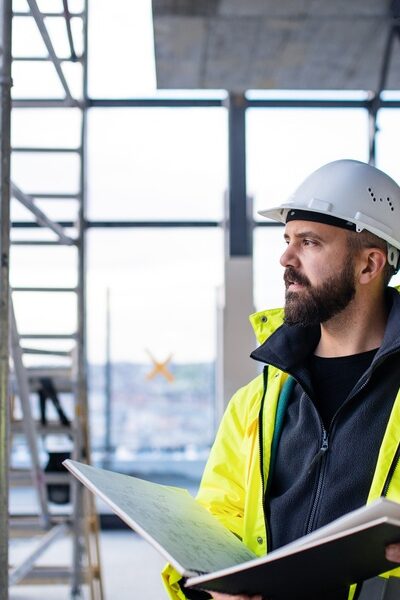 A man in a hard hat is holding a binder and is looking out into the distance while he’s standing in a construction zone.