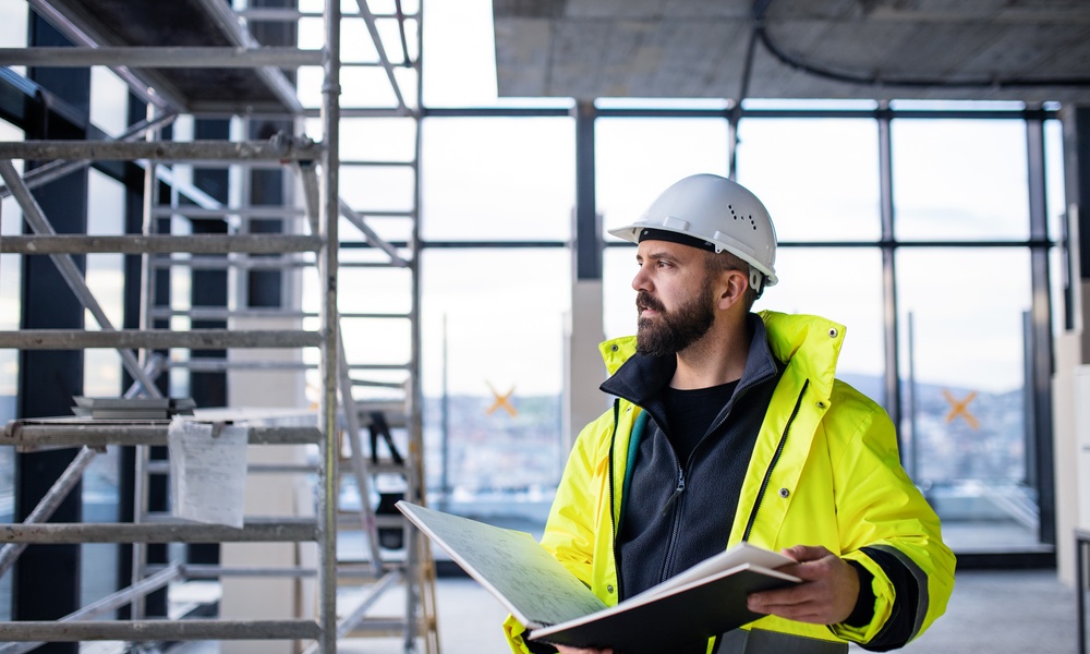 A man in a hard hat is holding a binder and is looking out into the distance while he’s standing in a construction zone.