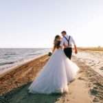 A bride and groom walking alone on a sandy beach on a sunny day. The ocean tide washes up onto the shore.