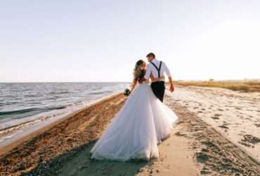 A bride and groom walking alone on a sandy beach on a sunny day. The ocean tide washes up onto the shore.