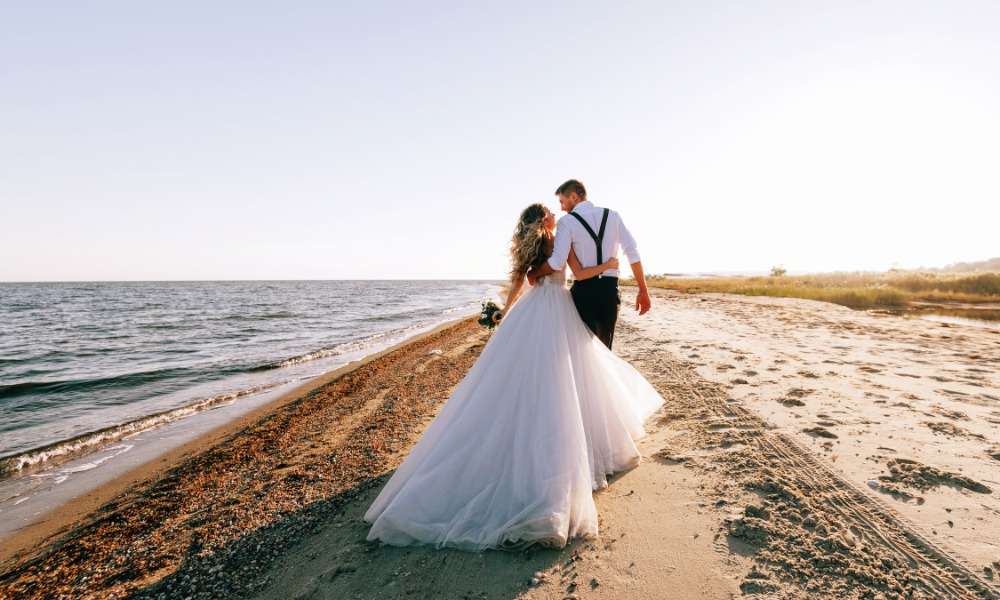 A bride and groom walking alone on a sandy beach on a sunny day. The ocean tide washes up onto the shore.