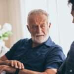 An older man is sitting on a couch next to a younger man. The older man has a mug in his left hand.