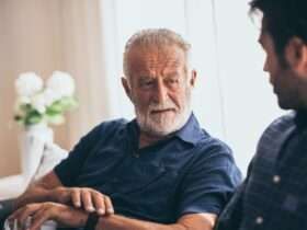An older man is sitting on a couch next to a younger man. The older man has a mug in his left hand.