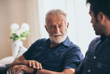 An older man is sitting on a couch next to a younger man. The older man has a mug in his left hand.