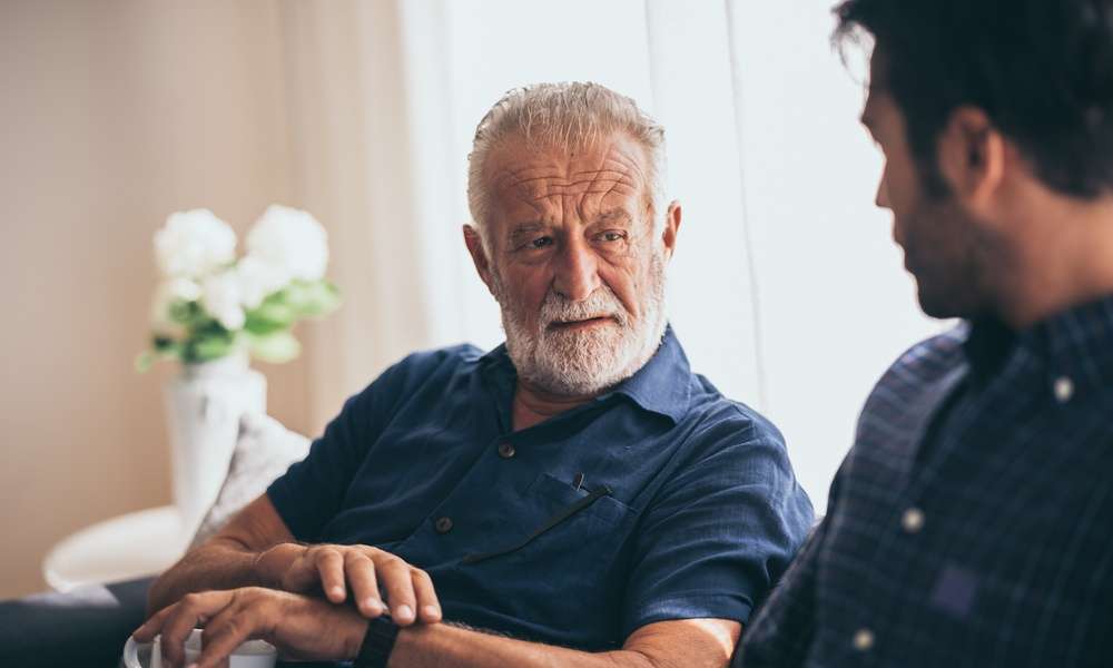 An older man is sitting on a couch next to a younger man. The older man has a mug in his left hand.