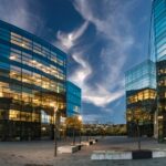 Modern glass office buildings with illuminated interiors surround an open plaza beneath a dramatic evening sky at dusk.