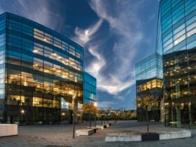 Modern glass office buildings with illuminated interiors surround an open plaza beneath a dramatic evening sky at dusk.