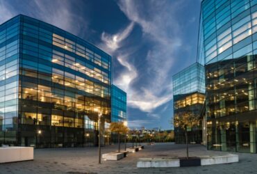 Modern glass office buildings with illuminated interiors surround an open plaza beneath a dramatic evening sky at dusk.