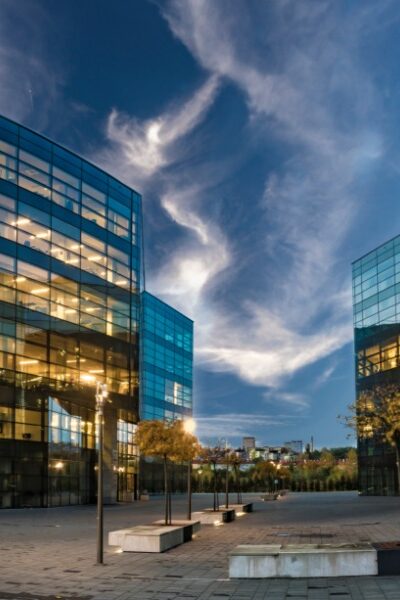 Modern glass office buildings with illuminated interiors surround an open plaza beneath a dramatic evening sky at dusk.