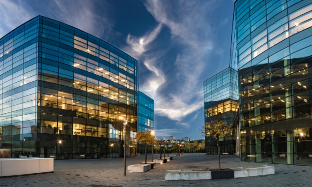 Modern glass office buildings with illuminated interiors surround an open plaza beneath a dramatic evening sky at dusk.
