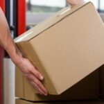 A person is kneeling over a pile of boxes as they pick one up. They’re holding a cardboard box with both hands.