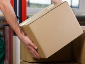 A person is kneeling over a pile of boxes as they pick one up. They’re holding a cardboard box with both hands.