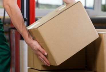 A person is kneeling over a pile of boxes as they pick one up. They’re holding a cardboard box with both hands.