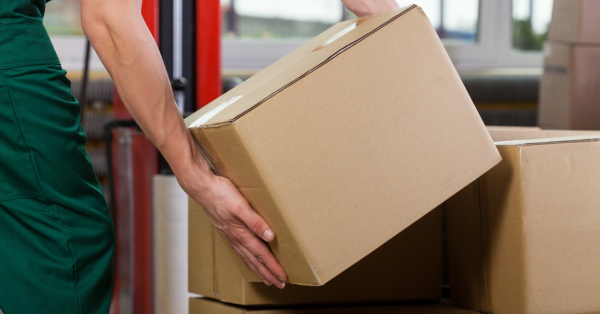 A person is kneeling over a pile of boxes as they pick one up. They’re holding a cardboard box with both hands.