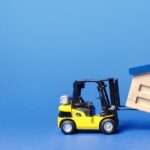 A yellow toy forklift lifts a wooden model of a factory. The factory has slanted blue roofing sections and a chimney.