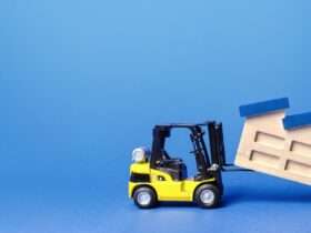 A yellow toy forklift lifts a wooden model of a factory. The factory has slanted blue roofing sections and a chimney.