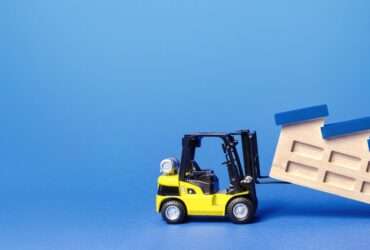 A yellow toy forklift lifts a wooden model of a factory. The factory has slanted blue roofing sections and a chimney.