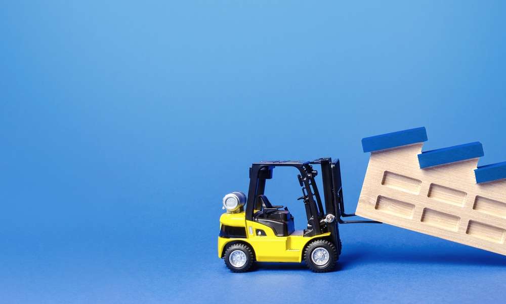 A yellow toy forklift lifts a wooden model of a factory. The factory has slanted blue roofing sections and a chimney.
