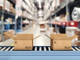 A close-up of a roller conveyor belt with multiple open cardboard boxes inside a warehouse with tall shelves.