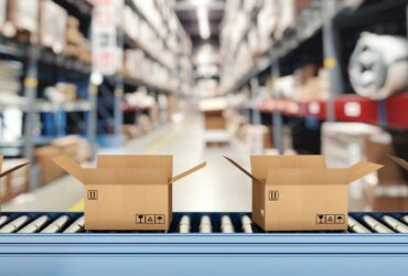 A close-up of a roller conveyor belt with multiple open cardboard boxes inside a warehouse with tall shelves.