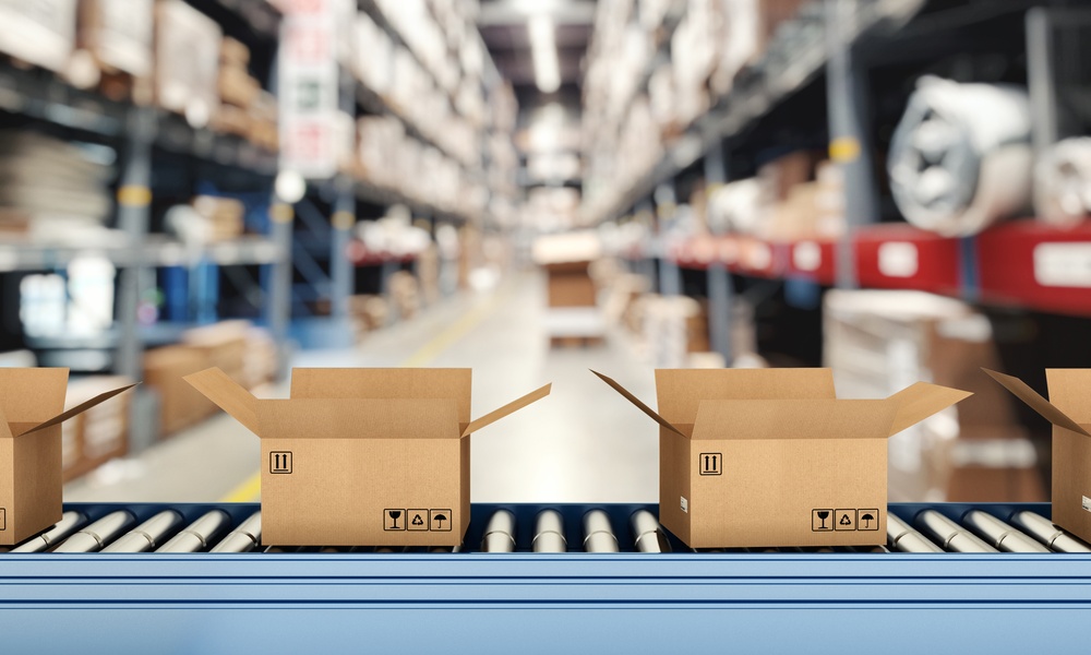 A close-up of a roller conveyor belt with multiple open cardboard boxes inside a warehouse with tall shelves.