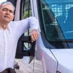 A man stands next to a delivery van and has his elbow on the window frame. He has glasses on the top of his head.