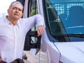 A man stands next to a delivery van and has his elbow on the window frame. He has glasses on the top of his head.