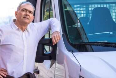 A man stands next to a delivery van and has his elbow on the window frame. He has glasses on the top of his head.