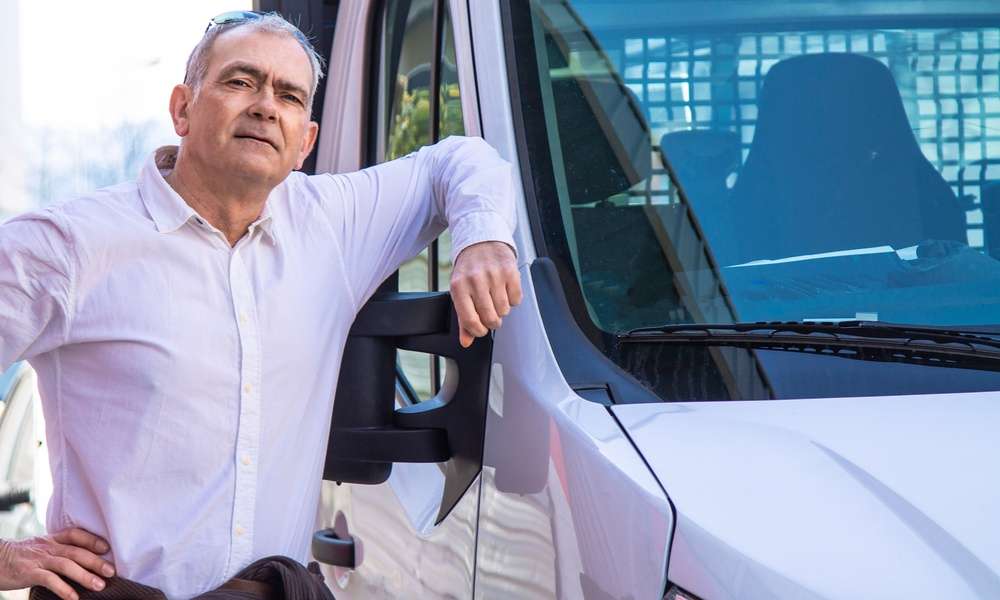 A man stands next to a delivery van and has his elbow on the window frame. He has glasses on the top of his head.