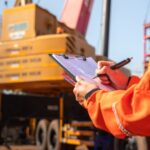 A close-up of a person with an orange work jacket writing with a pen on a clipboard with a large crane in the background.