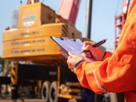 A close-up of a person with an orange work jacket writing with a pen on a clipboard with a large crane in the background.