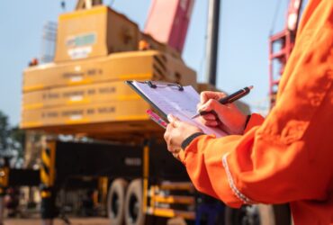 A close-up of a person with an orange work jacket writing with a pen on a clipboard with a large crane in the background.