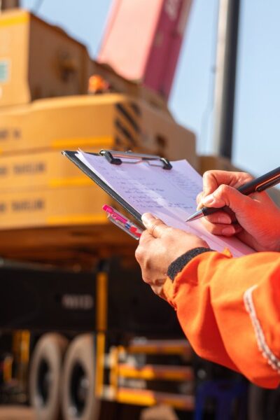 A close-up of a person with an orange work jacket writing with a pen on a clipboard with a large crane in the background.