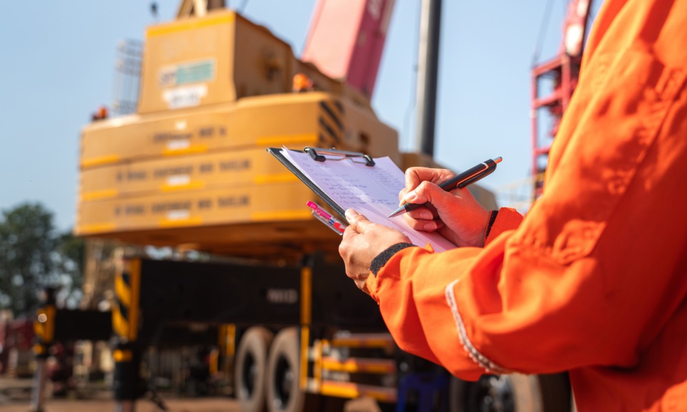 A close-up of a person with an orange work jacket writing with a pen on a clipboard with a large crane in the background.