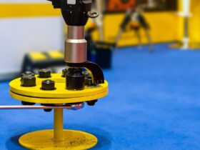 An electric torque wrench is tightening bolts on a yellow metal part with a silver wrench and a blue floor nearby.
