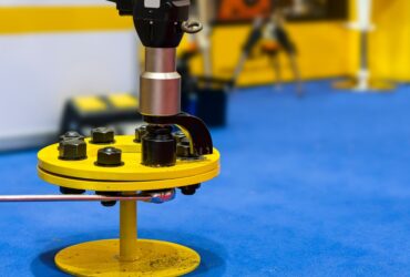An electric torque wrench is tightening bolts on a yellow metal part with a silver wrench and a blue floor nearby.