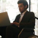 A man in a wheelchair wearing a black suit while working on a laptop on his lap inside a room with large windows.