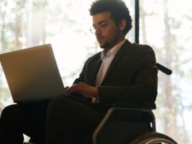 A man in a wheelchair wearing a black suit while working on a laptop on his lap inside a room with large windows.
