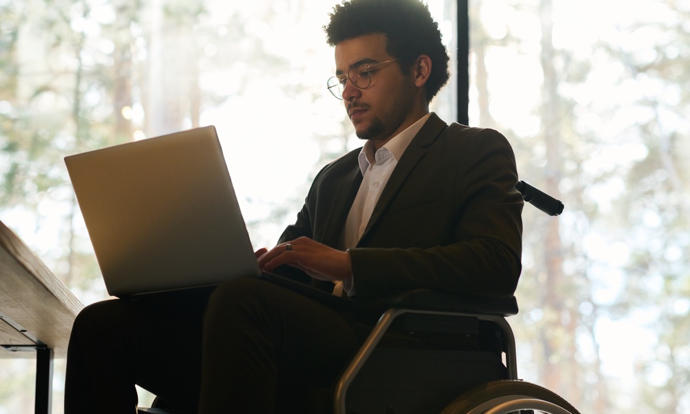 A man in a wheelchair wearing a black suit while working on a laptop on his lap inside a room with large windows.