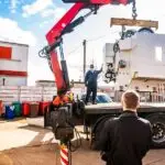 A crane truck lifts a large white industrial machine from a flatbed while workers stand nearby in an outdoor yard.