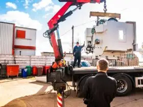 A crane truck lifts a large white industrial machine from a flatbed while workers stand nearby in an outdoor yard.
