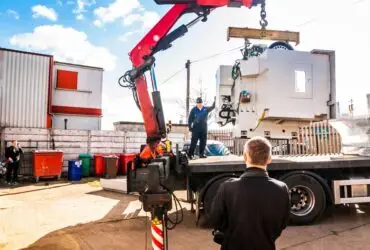 A crane truck lifts a large white industrial machine from a flatbed while workers stand nearby in an outdoor yard.