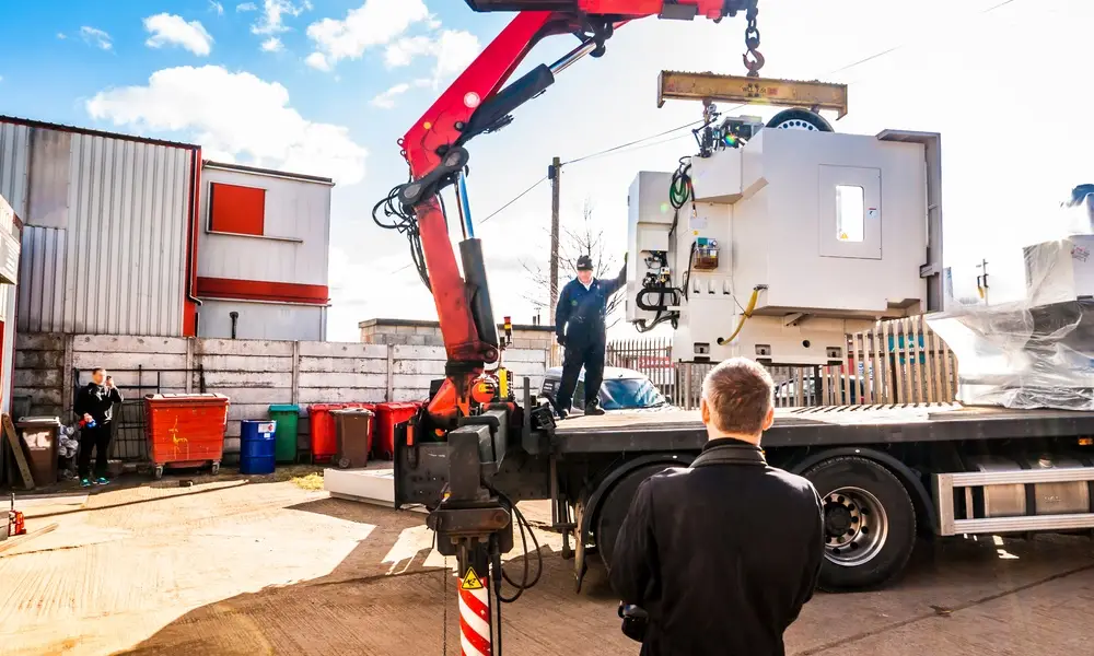 A crane truck lifts a large white industrial machine from a flatbed while workers stand nearby in an outdoor yard.