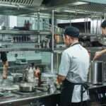 Three chefs working in a commercial kitchen. Steam rises from pots and pans cooking on the stovetops.
