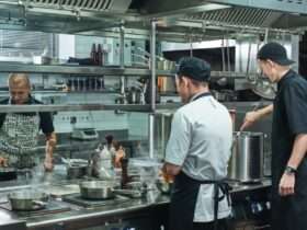 Three chefs working in a commercial kitchen. Steam rises from pots and pans cooking on the stovetops.