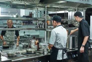 Three chefs working in a commercial kitchen. Steam rises from pots and pans cooking on the stovetops.