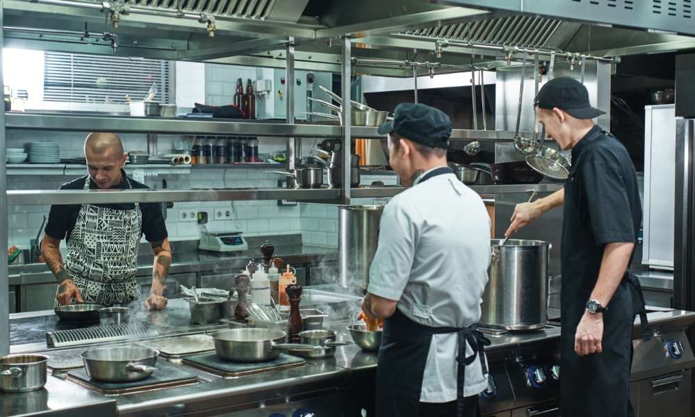 Three chefs working in a commercial kitchen. Steam rises from pots and pans cooking on the stovetops.
