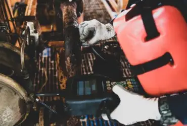 A worker inspects heavily corroded industrial pipes. An ultrasonic tool tests the remaining metal thickness.